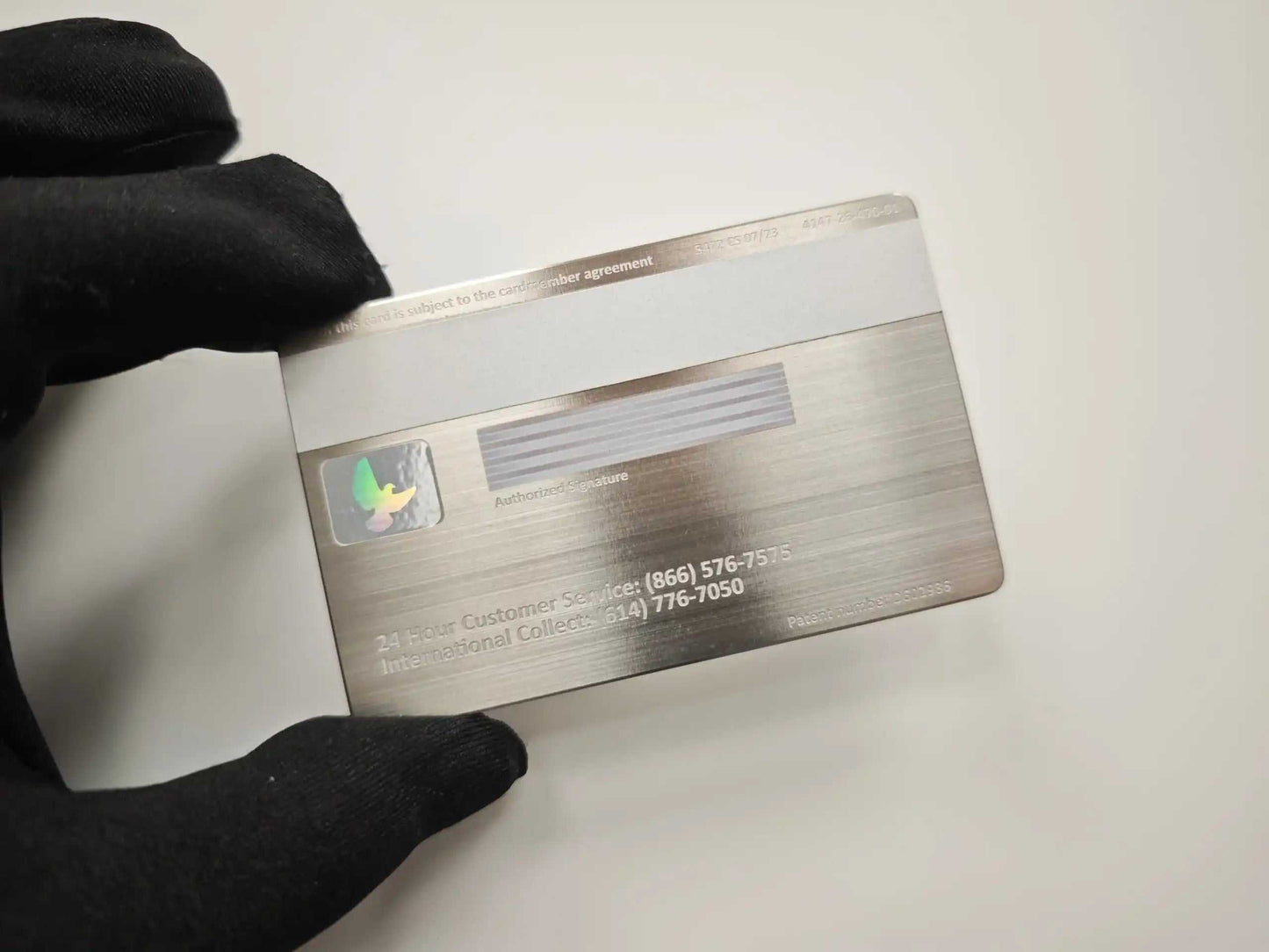 Brushed metal credit card with hologram, signature strip, held by gloved hand on white background.