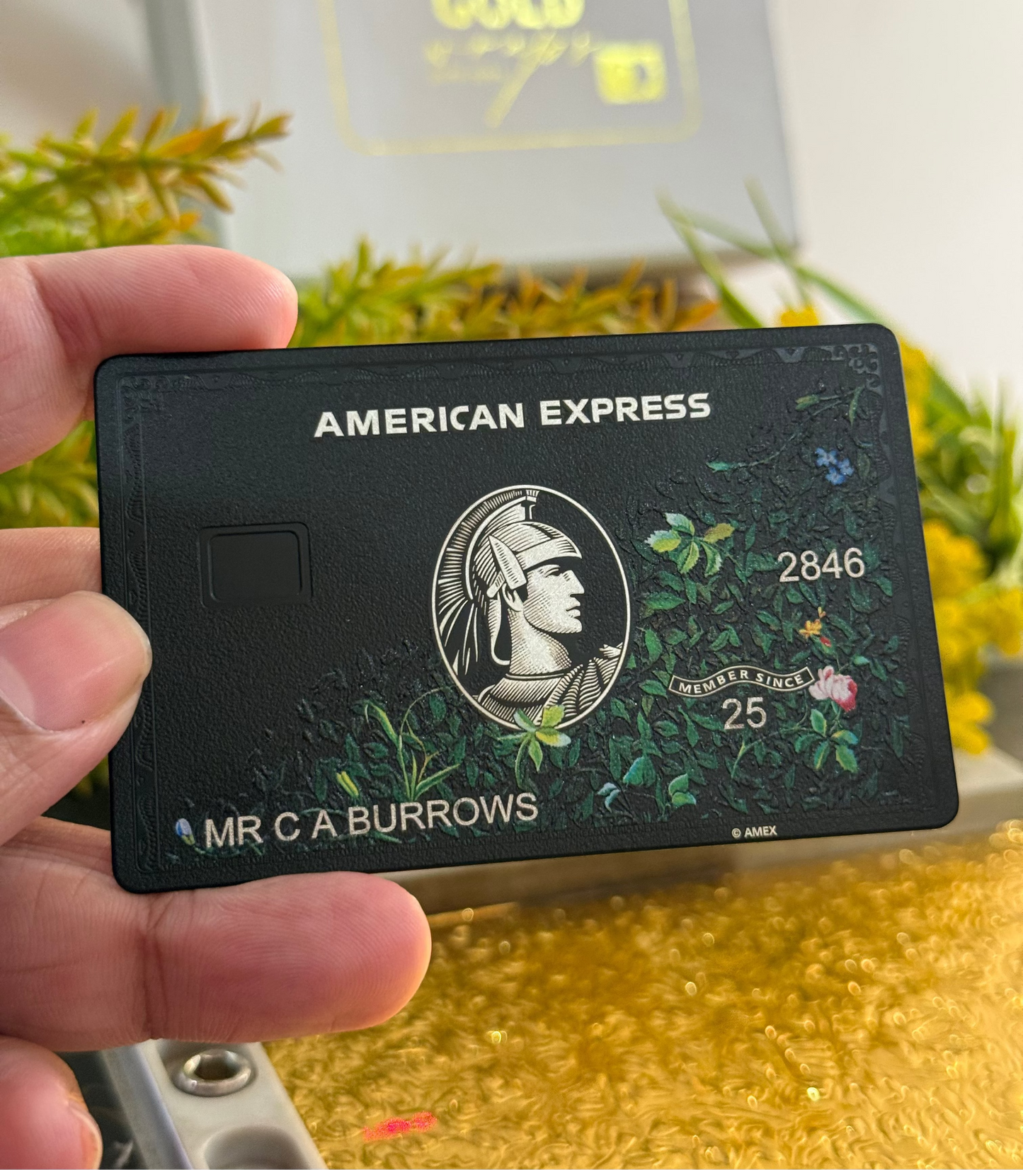 Hand holding black American Express custom metal card with floral design, gold background