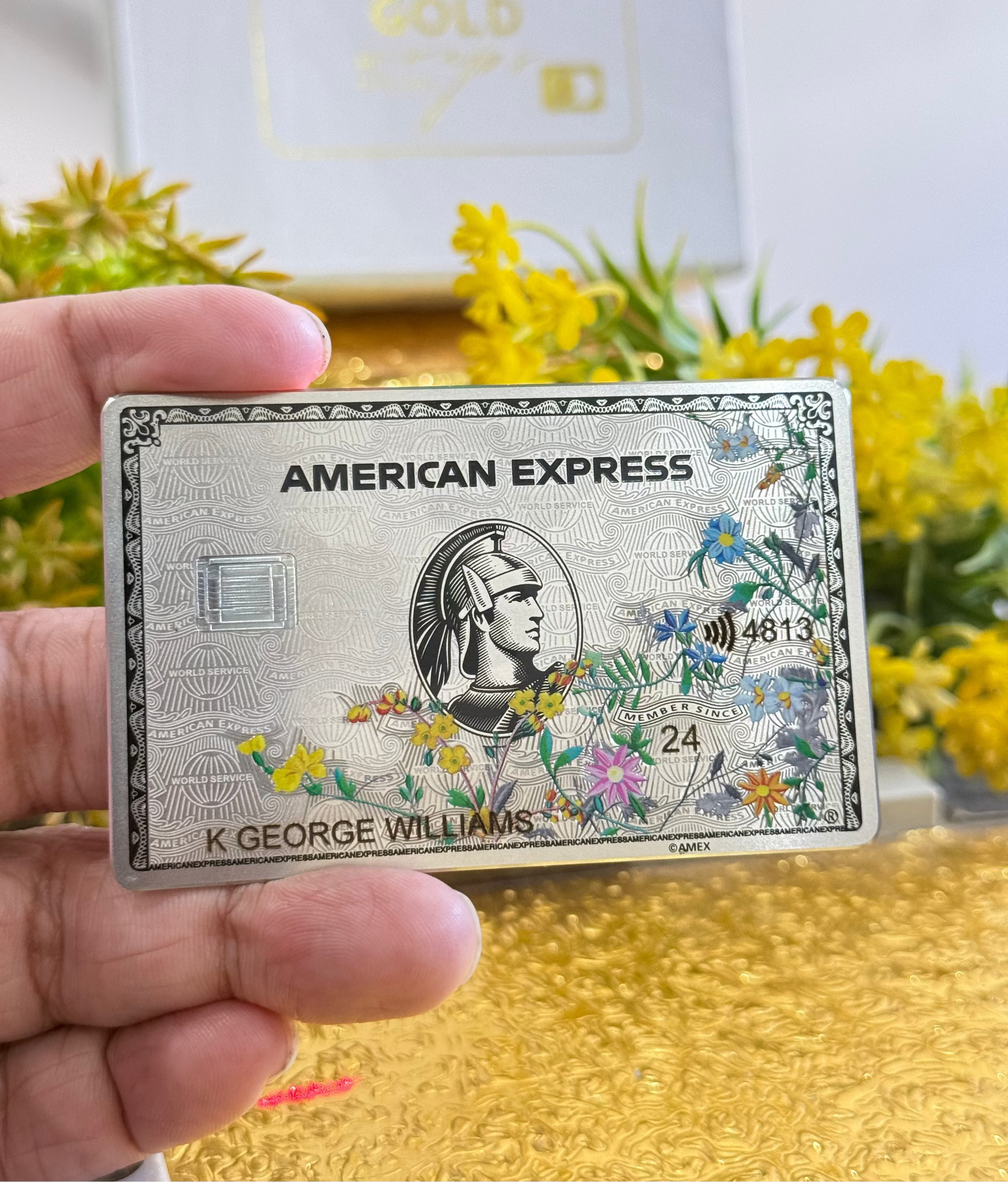 Custom metal American Express card with colorful floral design, held by hand, flowers in background