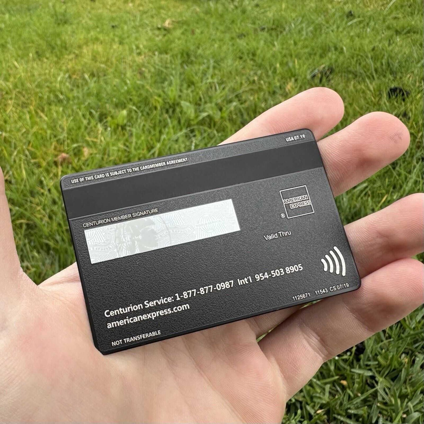 Hand holding a black metal American Express card outdoors on green grass